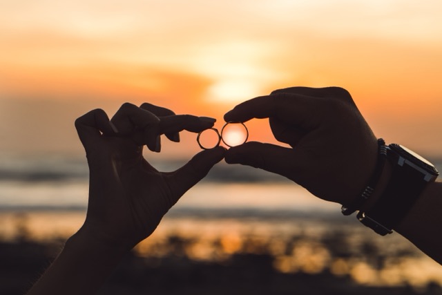 Two hands forming a heart around a pair of wedding rings at sunset