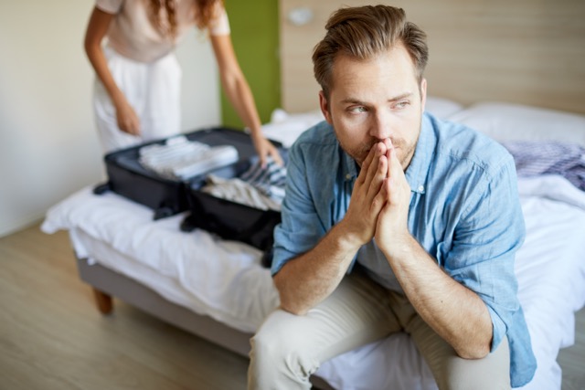 A man sitting on the edge of the bed, thinking, while his wife prepares to leave