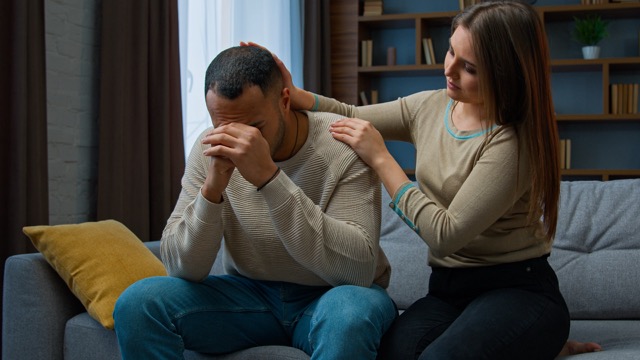Desperate sad man sitting on a sofa, upset, while his wife tries to support him