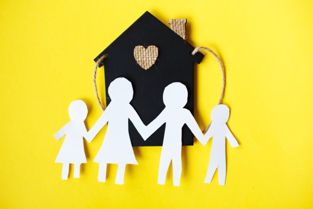 A paper cut family holding hands on a yellow background beside a small toy house
