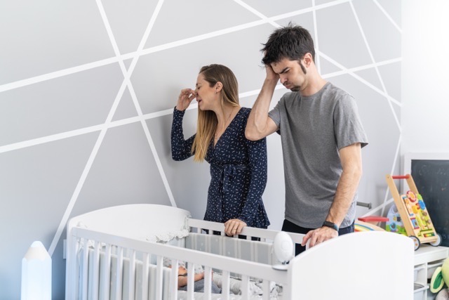A couple standing in a nursery beside a crib, both with their hands to their heads