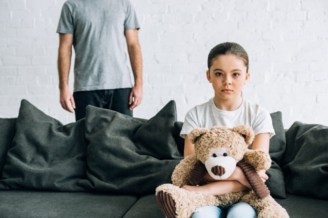 A father and his sad daughter, holding a teddy bear, sitting on a sofa