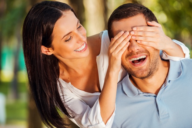 A woman covering her partner's eyes from behind, both smiling, outdoors
