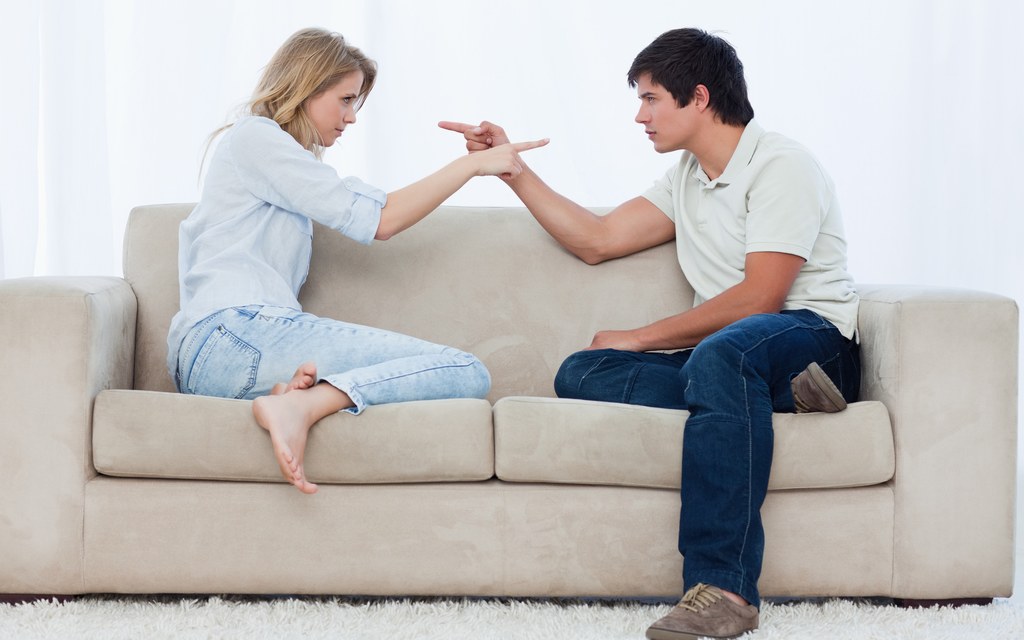 A couple sitting apart on a sofa mid argument, pointing at each other