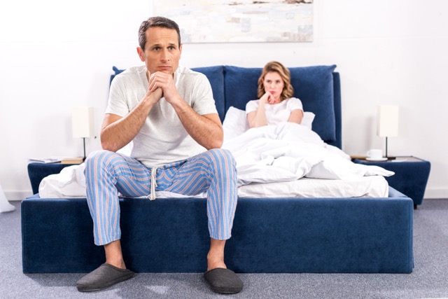 An upset man sitting on the bed with his wife behind him