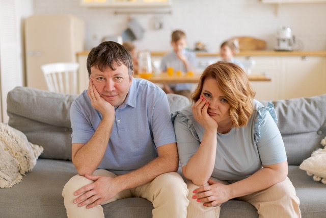 Tired parents sitting on the couch while their children run and shout in the kitchen