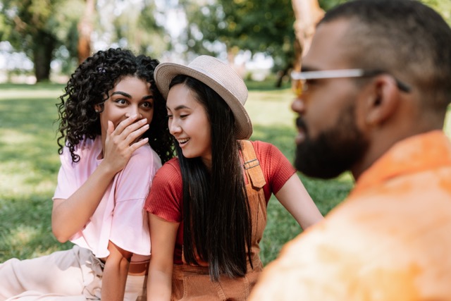 Two women relaxing at a picnic in the park while someone gossips