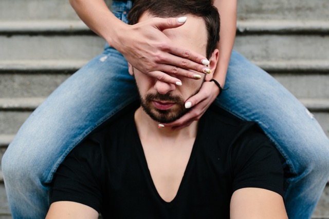 Catty behavior in a relationship: a man and woman against a gray wall, she is covering his face