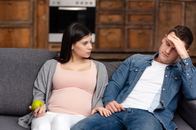 A pregnant woman holding an apple and looking at her husband on the sofa