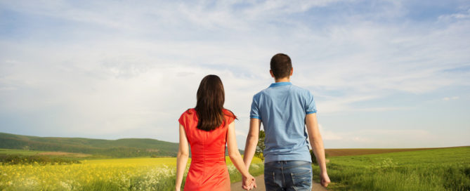 Man and woman holding hands in a field with a long dirt road ahead of them
