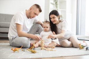 Benefits of Play for Kids: Loving caucasian parents in leisurewear sitting with small kid in white bodysuit and building toy tower from wooden blocks on floor of living room. Excited family bonding together using board game.