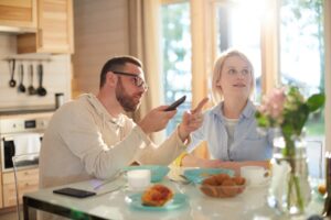 Encouraging Your Children:Young bearded Caucasian man pointing at TV while sitting at kitchen table with his wife, having breakfast and watching morning news attentively