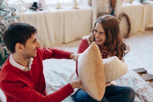 Save Marriage: Happy loving caucasian couple is getting crazy on the Christmas eve near the New Year tree, they are sitting on the bed, laughing a lot, fighting with pillows