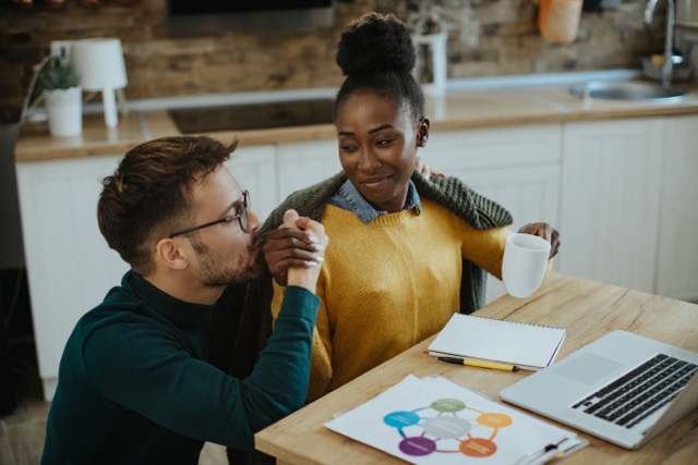 Save Marriage: Young African American woman drinking coffee and working on laptop while her husband is kissing her hand at home.
