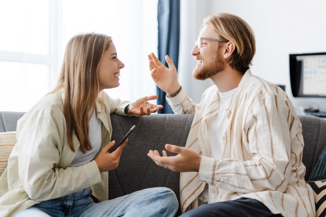 Young couple in a cozy living room, chatting and laughing on a sofa, radiating happiness and togetherness. Enjoying modern life with smartphones, they bond over their intimate connection
