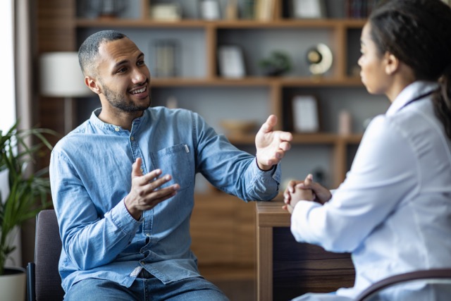 Handsome african american young man having appointment with female doctor, looking at black woman therapist, gesturing and smiling, modern clinic interior, copy space