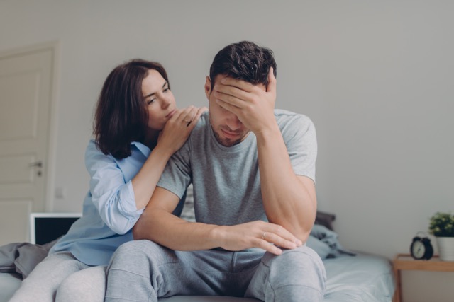 Miserable Husband Syndrome: Attractive female consoles her sad boyfriend who has depression and some problems, pose at bedroom on bed, sit against domestic interior. Wife and husband try solve difficult situation in life.