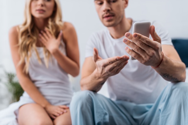 Signs of Infidelity In A Relationship: cropped shot of emotional man holding smartphone and scared woman sitting behind on bed, mistrust