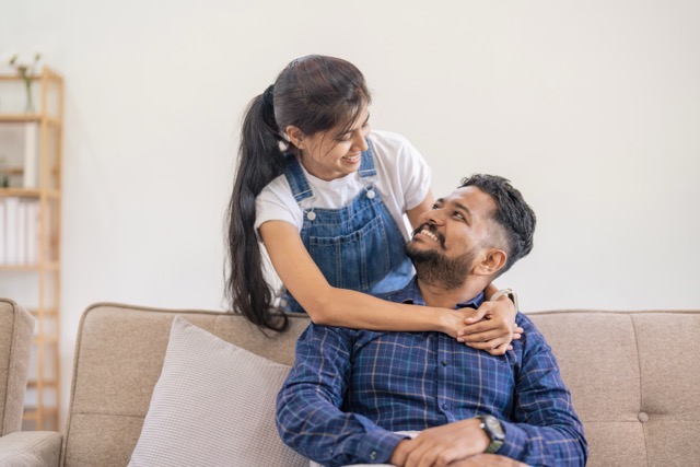 Miserable Husband Syndrome: Happy lovely young Indian couple together at home, young wife hugging from behind her husband, sitting and rest on sofa in home, portrait of romantic multiracial couple in love.
