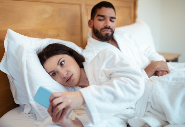 Signs of Infidelity In A Relationship: Jealousy Problem. Husband Looking At Cheating Wife When Woman Texting On Smartphone Communicating Online Lying In Bedroom At Home. Jealous Boyfriend Suspecting Affair. Selective Focus