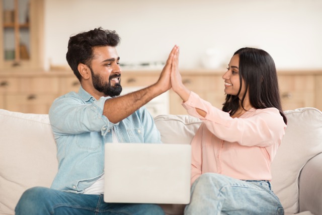 Joyful indian couple using laptop computer and giving high-five celebrating success or great news, sitting on sofa at home. Technology and internet