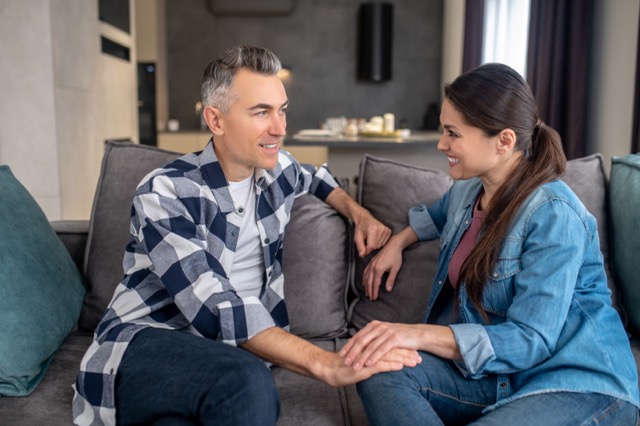 Smiling middle aged man in casual clothes listening to touching happy shining woman looking at each other sitting on sofa at home