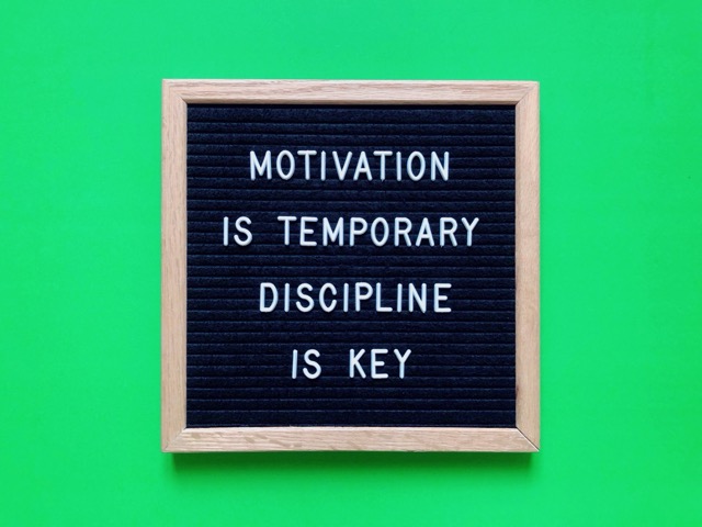 The board has a black background with white letters and a wooden frame, set against a bright green background. It conveys a powerful message about the importance of discipline over fleeting motivation.