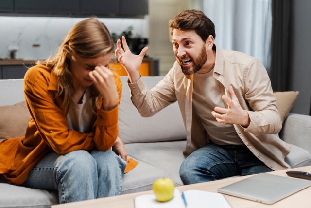 Portrait of upset woman crying, being yelled at by angry, abusive man, sitting on sofa in room at home. Concept of quarrel, conflict