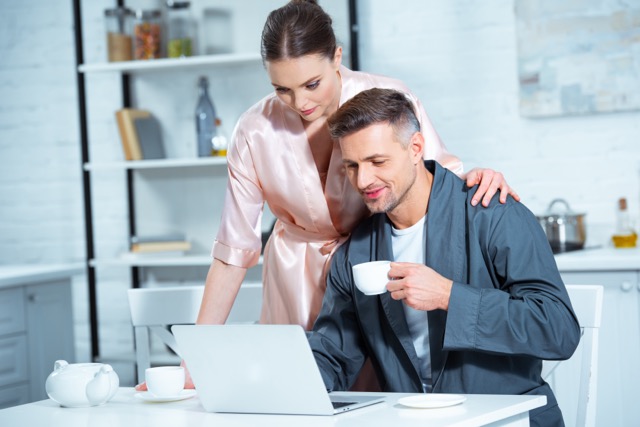 Passive aggression in marriage: selective focus of beautiful adult couple in robes using laptop during breakfast in kitchen