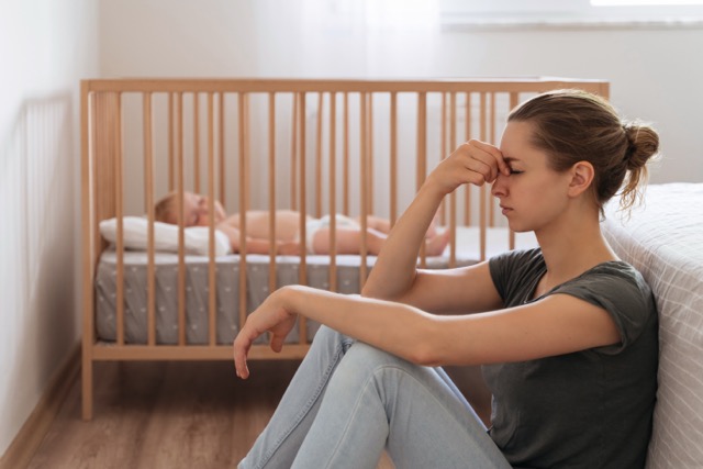 Divorcing With A Baby: young divorced mother sitting on floor in child's bedroom while baby sleeps in bed, trying to calm down, suffering postnatal depression symptoms