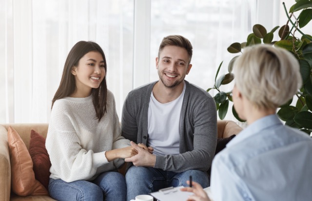 Personal Coaches In Miami: Reconciliation. Smiling Multiethnic Spouses Holding Hands Reconciling After Quarrel Sitting In Family Psychotherapist's Office
