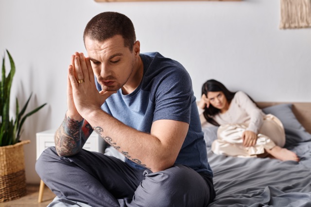 Miserable Husband Syndrome: young depressed tattooed man sitting on bed near offended asian wife, family divorce concept