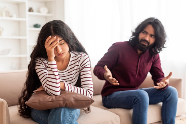 Feeling Smothered in Relationships: Breaking Up And Divorce Concept. Young married indian couple having fight, guy yelling at crying lady, gesturing at home. Furious man shouting at his wife or girlfriend, sitting on sofa at living room