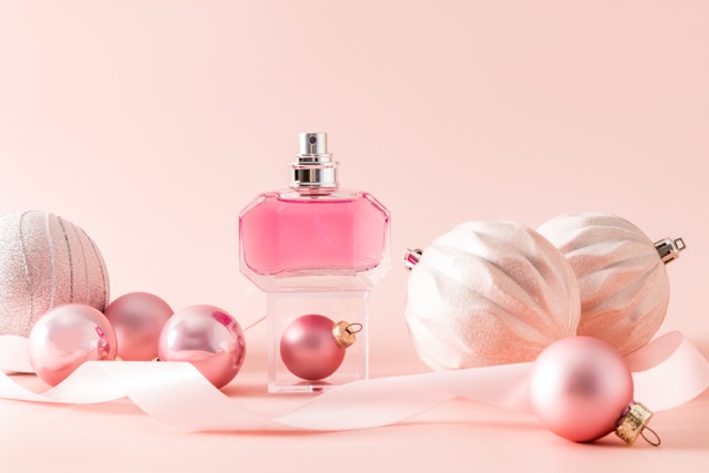 A chic bottle of women's perfume stands on a transparent cube podium among pink New Year's balls. Blank bottle mockup. Presentation of the fragrance.