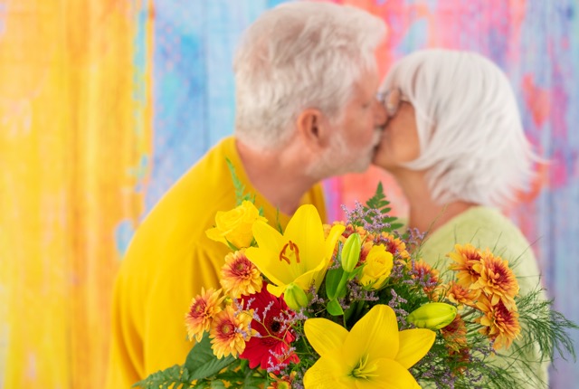 Blurred senior couple kissing on colorful background holding a flower bouquet. Romantic portrait of elderly caucasian couple. Woman receives flowers as present from husband