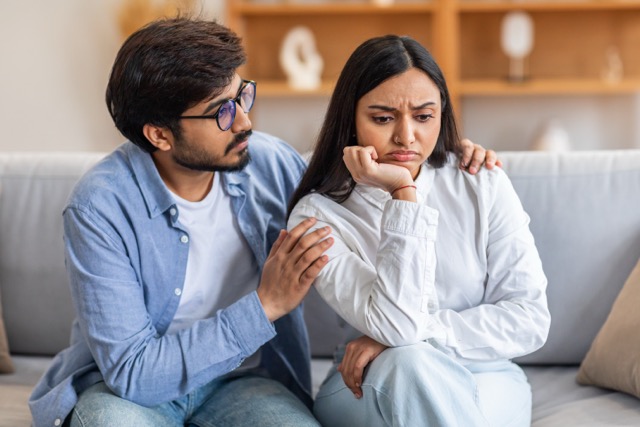 man with glasses gently consoles a distressed woman sitting beside him on a couch in a comfortable, well-lit living room.
