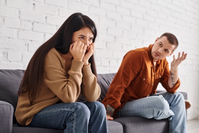 discouraged man talking to offended asian wife sitting on couch in living room, divorce concept