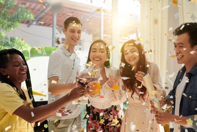 Group of young excited intercultural friends clinking with red wine in wineglasses and orange juice while enjoying home party with confetti