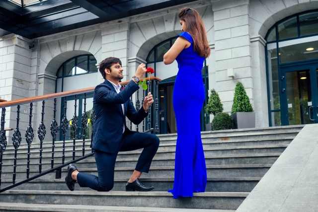 happy asian man in stylish black suit falling knee in front of his beautiful woman in blue long dress asking proposal and giving gold ring on the background of the restaurant street.