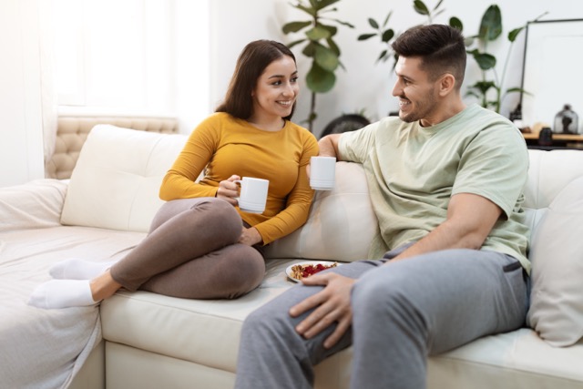 Happy attractive millennial couple drinking coffee at home. Cheeful young man and woman wearing comfy homewear sitting on couch in living-room, drinking tea and have conversation, copy space