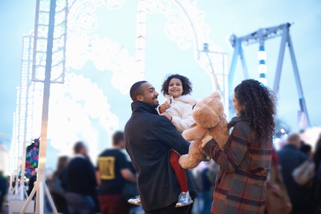 Mother and father in amusement park carrying smiling girl and teddy bear