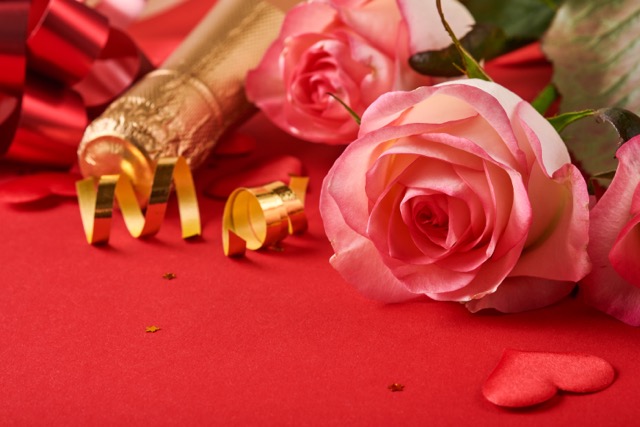 Rose Day Gift Ideas Beyond Flowers: Pink roses flowers, champagne, gift, golden ribbons and confetti red hearts on red background. Top view flat lay with space for your greetings. Valentines day background and greeting card.