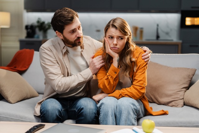 Couple portrait, bearded man comforting beautiful young woman, she has toothache, sitting on sofa at home, hugging her. Relationship concept