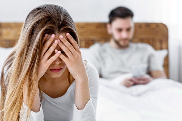  Focus on sad woman, covering her face with hands, sitting on bed, man typing on smartphone in minimal bedroom interior, free space.