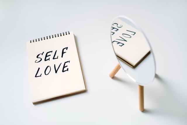 Self love, state of appreciation for oneself, physical, psychological and spiritual growth, taking care of own needs, wellbeing. Notepad with text Self love and reflection in the mirror.