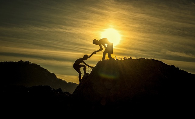 Two people silhouetted against a sunset, with one person helping the other climb a rock, symbolizing teamwork and support.