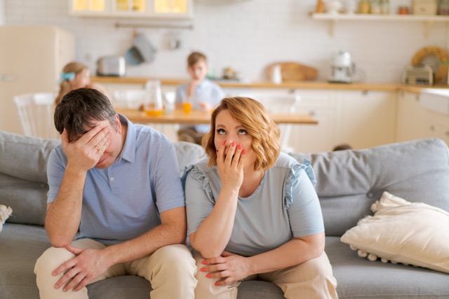 Tense parents sitting on sofa tired crazy loud hyperactive little kids, siblings at home. An unhappy married couple is annoyed by a naughty little daughter and two sons with headaches.