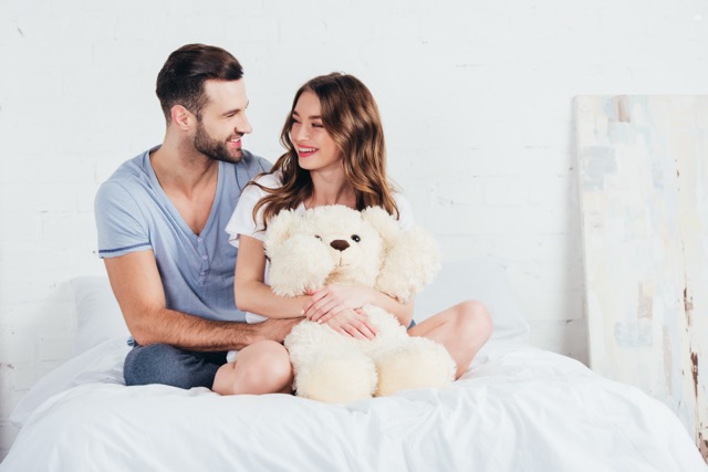 Young couple hugging teddy bear while sitting on bed with white bedding